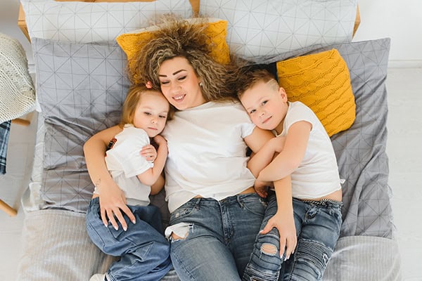 mom-with-kids Happy mother with two children lying on bed, family moments, cozy home scene.
