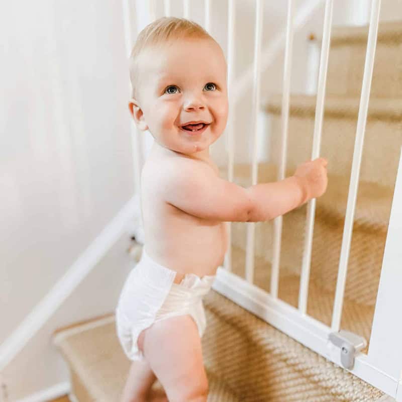 A smiling baby standing near a safety gate on a staircase, indoor home setting, Ocean City Moms.