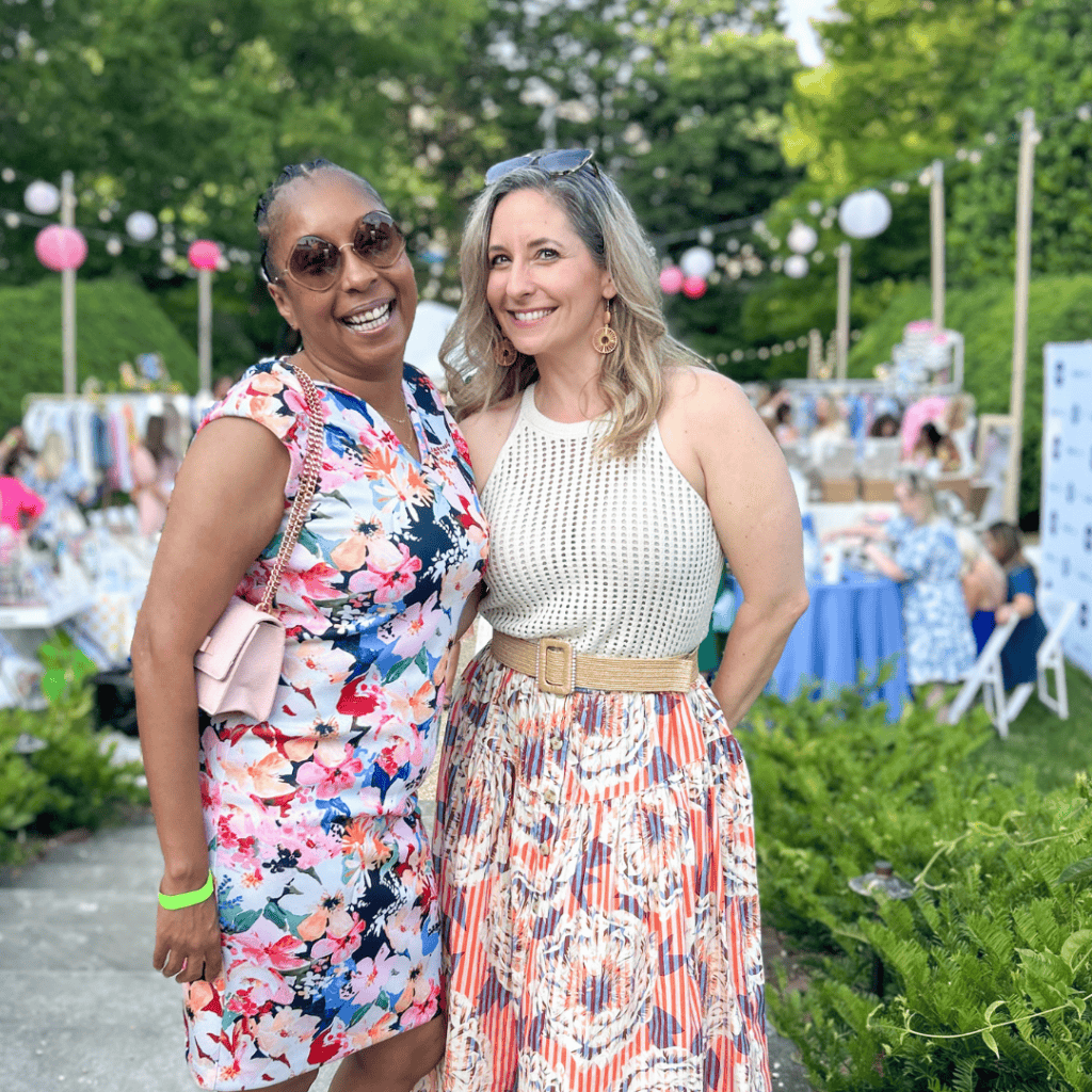 Two women smiling at an outdoor event with colorful decorations and tables in the background.
