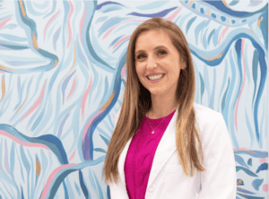 Smiling woman in white blazer and pink top standing in front of colorful abstract mural.