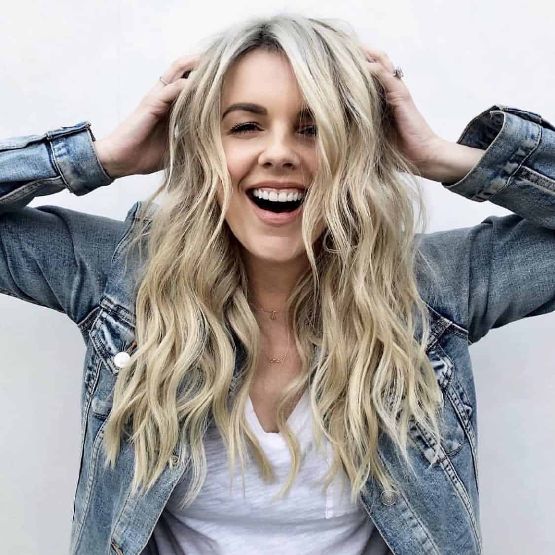 Happy woman with long blonde hair wearing denim jacket smiling and holding her hair.