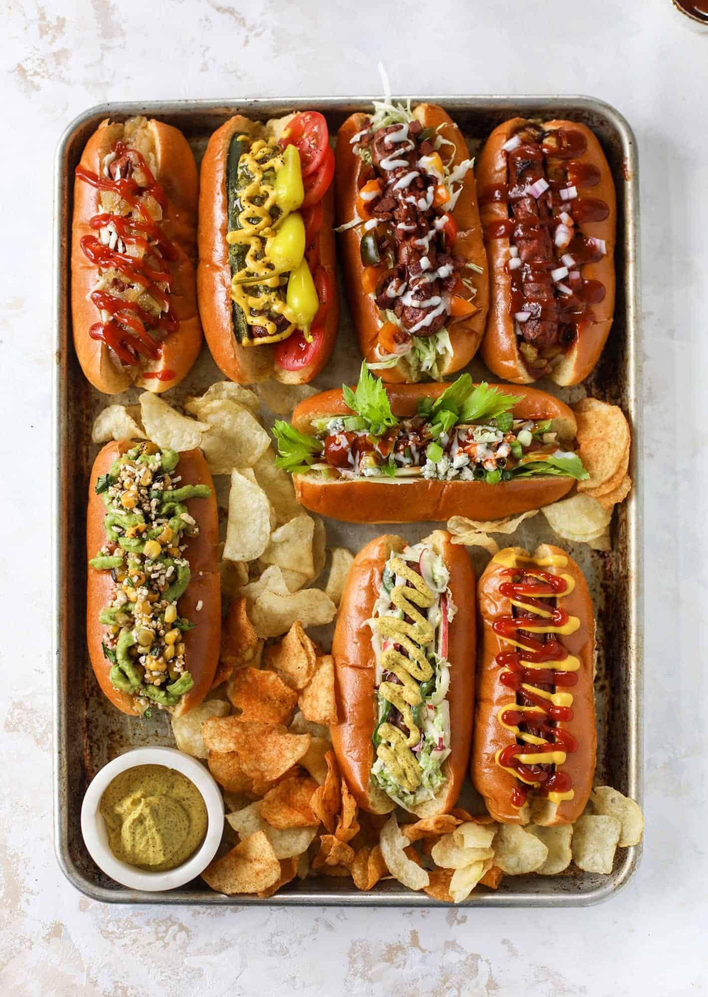 Assorted gourmet hot dogs with toppings on a metal tray with chips and mustard.