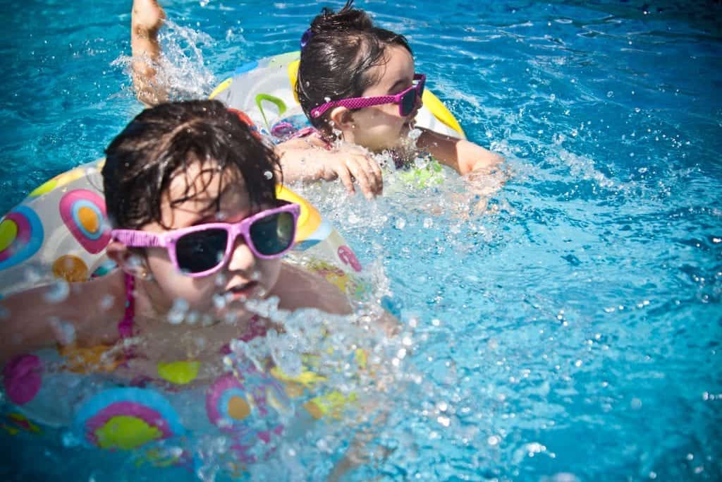 Two children swimming in a pool with colorful floaties and pink sunglasses.